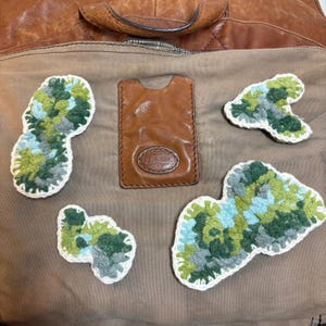 May include: A brown leather card holder with a Fossil logo embossed on the front, surrounded by four irregular, textured crochet patches in shades of forest green, lime green, light blue, and grey, each outlined with a white crocheted border.