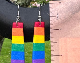 Puede incluir: Un par de pendientes hechos a mano con los colores del arcoíris. Cada pendiente tiene forma rectangular, con rayas rojas, naranjas, amarillas, verdes, azules y moradas. Los pendientes miden unos 5 cm de largo.
