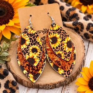 May include: A pair of layered teardrop-shaped earrings featuring a sunflower and leopard print pattern on the top layer, a middle layer of copper-coloured glitter, and a bottom layer with a sunflower and leopard print design on silver hooks.