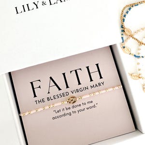 May include: A bracelet with pink beads and gold accents is displayed inside a white box. The card inside the box reads "FAITH THE BLESSED VIRGIN MARY" with the quote "Let it be done to me according to your word."