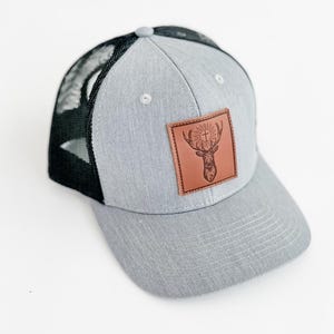 May include: A gray and black trucker hat with a brown leather patch featuring a deer with antlers and a cross.