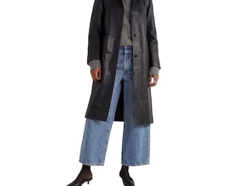 May include: A black leather trench coat worn over a grey knit sweater and light blue wide-leg denim jeans. The coat features a classic collar, long sleeves, and a button-front closure. The outfit is completed with black pointed-toe kitten heels.