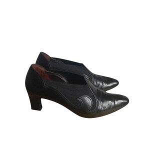 Puede incluir: Un par de zapatos de cuero negro con tacón bajo. Los zapatos tienen punta afilada y un diseño decorativo en el lateral. El tacón mide aproximadamente 5 cm de alto.