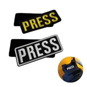 May include: Two rectangular patches with the word "PRESS" in bold letters. One patch has yellow text on a black background, and the other has white text on a black background. Both patches have a black backing.