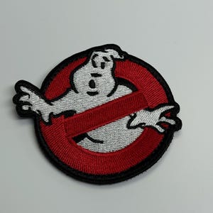 May include: Embroidered patch featuring the Ghostbusters logo. The design includes a white ghost figure inside a red circle with a red slash through it. The patch has a black border.