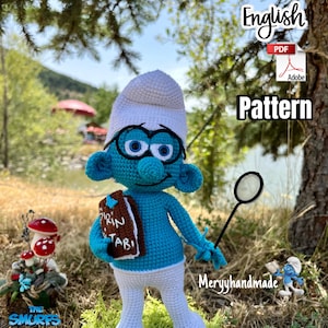 Könnte beinhalten: Eine gehäkelte Schlumpf-Puppe mit einem weißen Hut und einer Brille, die ein Buch und eine Lupe hält. Die Puppe ist blau mit einem weißen Hemd und einer Hose. Die Puppe steht auf einer grünen Oberfläche mit einem Pilz und einem Schild, auf dem "Die Schlümpfe" steht.