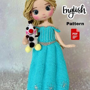 May include: A handmade crochet doll in a turquoise dress, holding a small snowman. The doll has blonde hair, blue eyes, and a silver tiara. The dress is detailed with silver accents and snowflake patterns. The image includes the text "English Pattern PDF".