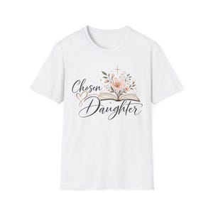 Puede incluir: Una camiseta blanca de manga corta con un estampado gráfico de un libro abierto del que brotan flores rosas y hojas verdes. El texto "Chosen Daughter" aparece en caligrafía negra debajo del libro, junto con el icono de un pequeño corazón.