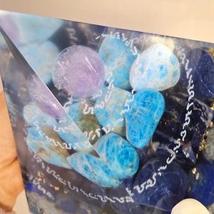 Pode incluir: Uma peça de arte em resina em forma de pirâmide preenchida com várias pedras preciosas azuis e roxas. As pedras variam em tonalidade, do azul claro ao índigo profundo, com algumas pedras de cor lavanda. A escrita branca é visível.