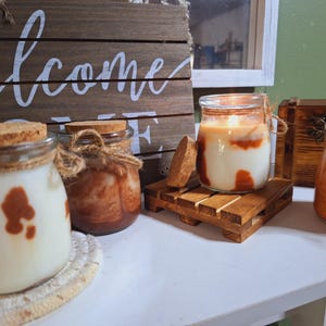 Peut inclure: Plusieurs bougies en pot de verre avec des couvercles en liège et des accents de ficelle sont présentées. Les bougies ont un aspect en couches avec de la cire blanche et marron. Un panneau en bois avec le mot "welcome" est en arrière-plan.