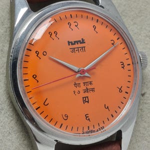 May include: A vintage wristwatch with a bright orange face and a silver-colored case. The watch face features black numerals and text in a foreign language. The watch has silver hands and a brown leather strap.