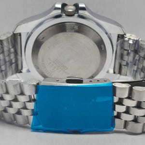 May include: Close-up of a silver-toned wristwatch with a stainless steel band. The watch face is visible, and the band is partially wrapped. The clasp is covered in blue protective film. The watch is a Seiko.
