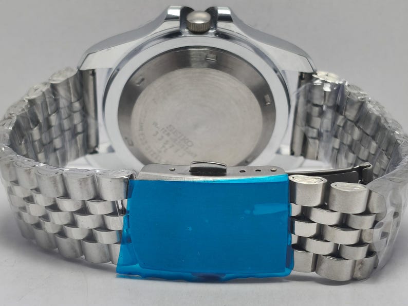 May include: Close-up of a silver-toned wristwatch with a stainless steel band. The watch face is visible, and the band is partially wrapped. The clasp is covered in blue protective film. The watch is a Seiko.