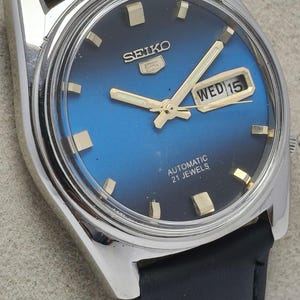 May include: A Seiko 5 automatic wristwatch with a blue and black gradient dial, gold-tone hour markers, and gold-tone hands. The watch features a day-date window at the three o'clock position and is fitted with a black leather strap.