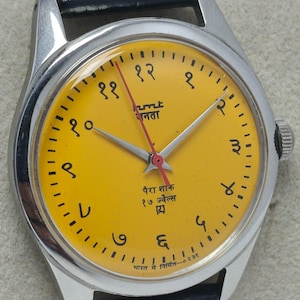 May include: A vintage wristwatch with a bright yellow face and silver-tone case. The watch features black numerals, a red second hand, and a black leather strap. The watch face includes text in a foreign language.