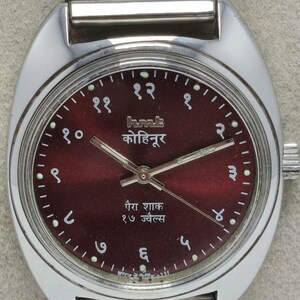 May include: A silver-toned wristwatch with a burgundy dial. The watch face features Arabic numerals and text in a foreign language. The watch has a metal band and a crown on the side.