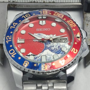 May include: A Seiko wristwatch with a red and blue bezel. The watch face features a wave design and the day of the week. The watch has a stainless steel band and a silver case. The bezel has numbers from 2 to 24.