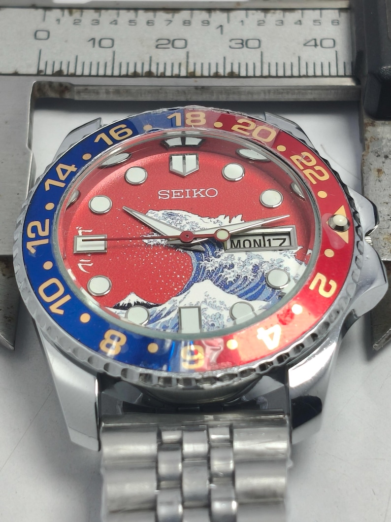 May include: A Seiko wristwatch with a red and blue bezel. The watch face features a wave design and the day of the week. The watch has a stainless steel band and a silver case. The bezel has numbers from 2 to 24.