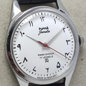 May include: A silver wristwatch with a white face, black Arabic numerals, and a red second hand. The watch face displays the text "hmk Janata PARA SHOCK 17 JEWELS MADE IN INDIA". The watch has a dark blue leather strap.
