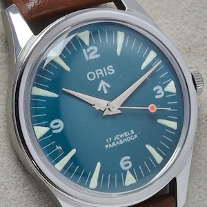 Peut inclure: Une montre ORIS vintage avec un cadran bleu sarcelle et un bracelet en cuir marron. La montre a des index et des aiguilles blancs, avec le chiffre 12 en haut. Le cadran affiche également le texte "17 JEWELS PARASHOCK".