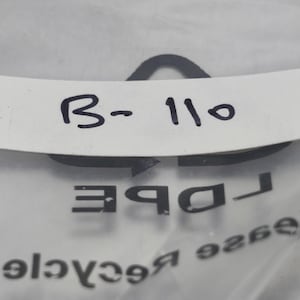 May include: A close-up shot of a white label with the text "B-110" in black. The label is partially obscured by a clear plastic bag with the word "DBE" and other text visible.