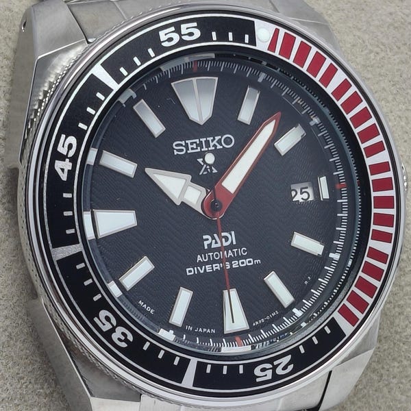 May include: A stainless steel Seiko PADI automatic diver's watch with a black dial and a date display. The watch features white and red accents on the bezel and hands. The watch is water resistant to 200m.