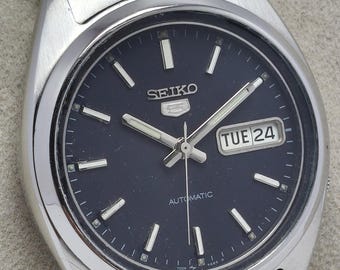 May include: A silver-toned Seiko 5 automatic wristwatch with a dark blue dial. The watch features a day and date display, luminous hands and markers, and a stainless steel band. The watch face displays the day as "TUE 24".