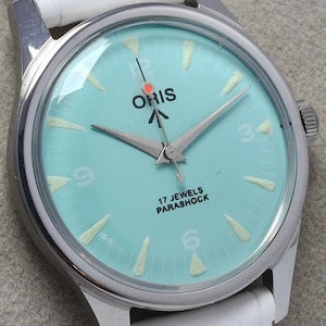 Puede incluir: Un reloj Oris vintage con esfera azul claro, manecillas plateadas y correa de cuero blanco. La esfera del reloj presenta el texto "17 Jewels Parashock". El reloj tiene una caja plateada y una correa de cuero blanco.