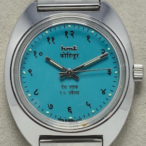 May include: A silver-toned wristwatch with a turquoise face. The watch face features Arabic numerals and text in a foreign script. The watch has a silver band and a crown on the side. The watch is a classic style.