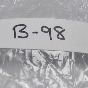 以下が含まれることがあります： 手書きの「B-98」という黒インクの文字が書かれた白いラベルのクローズアップ。ラベルは、しわのある外観の、質感のある反射面の上に配置されています。