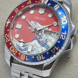 May include: A stainless steel Seiko watch with a red dial featuring a wave design and the text "SEIKO". The bezel is red and blue with gold numerals. The watch shows the day and date.