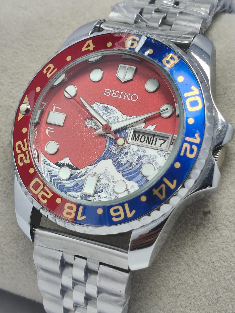 May include: A stainless steel Seiko watch with a red dial featuring a wave design and the text "SEIKO". The bezel is red and blue with gold numerals. The watch shows the day and date.