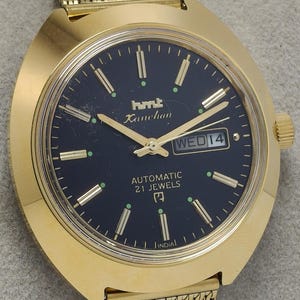 Op de afbeelding: Goudkleurig polshorloge met een donkerblauwe wijzerplaat. Het horloge heeft gouden wijzers en markeringen, een datumweergave en de tekst "hmt Kanchan" en "AUTOMATIC 21 JEWELS". Het horloge heeft een gouden mesh band.
