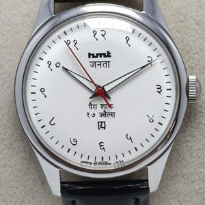 May include: A silver-toned wristwatch with a white face and black leather band. The watch face features black numerals, black and red hands, and text in a foreign language. The watch has a round face and a silver-toned bezel.