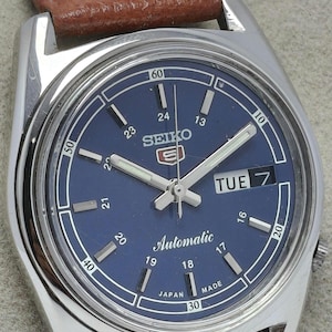 May include: A Seiko 5 automatic wristwatch with a round silver-tone stainless steel case and a blue dial. The dial features white hour markers, luminous hands, a day-date window at the three o'clock position, and a brown leather strap with stitching.