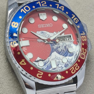 May include: A stainless steel Seiko watch with a red dial featuring a wave design and the word "SEIKO". The bezel is blue and red with gold numerals. The watch band is silver.