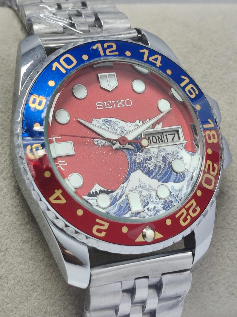 May include: A stainless steel Seiko watch with a red dial featuring a wave design and the word "SEIKO". The bezel is blue and red with gold numerals. The watch band is silver.