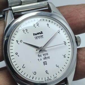 May include: A silver wristwatch with a white face and black numerals. The watch has a silver band and features text in a foreign language. The watch hands are black and the watch has a silver crown.