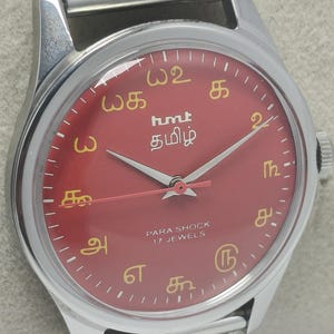 May include: A wristwatch with a red face and silver case and strap. The watch face has gold markings and the text "hmt தமிழ்" and "PARA SHOCK 17 JEWELS". The strap is a mesh design.