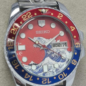 May include: A stainless steel Seiko watch with a red and blue bezel. The watch face features a red background with a white and blue wave design, a mountain, and the day of the week. The watch has a silver band.