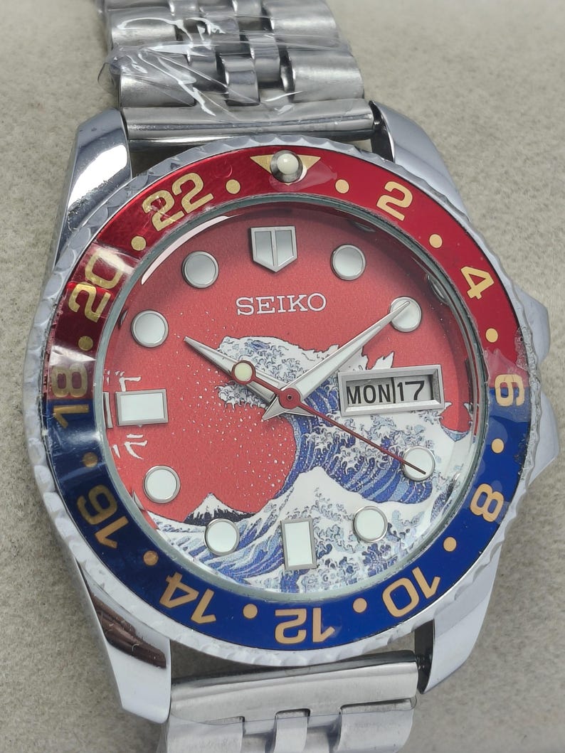 May include: A stainless steel Seiko watch with a red and blue bezel. The watch face features a red background with a white and blue wave design, a mountain, and the day of the week. The watch has a silver band.