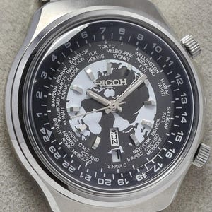 May include: A silver-toned wristwatch with a black dial featuring a world map design and city names. The watch face includes the brand name "RICOH" and displays the day and date. The watch has a stainless steel band.