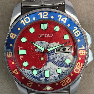 May include: A stainless steel Seiko watch with a red dial featuring a wave design and luminous markers. The bezel is blue and red with white numerals. The watch band is also stainless steel.