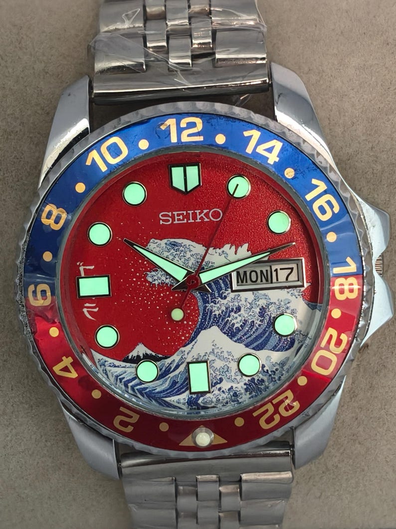 May include: A stainless steel Seiko watch with a red dial featuring a wave design and luminous markers. The bezel is blue and red with white numerals. The watch band is also stainless steel.