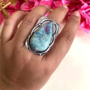 Könnte beinhalten: Ein silberner Ring mit einem großen, tropfenförmigen, grün, blau und rosa gefärbten Edelstein, der in einem dekorativen Silberrahmen gefasst ist.