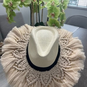May include: A cream-colored fedora hat with a woven straw crown and a black band. The hat features a macrame design around the brim with long fringe. The hat is a stylish accessory.