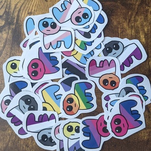 May include: A collection of colorful stickers featuring cartoon characters with different pride flag designs. The stickers are printed on white paper and have a glossy finish.
