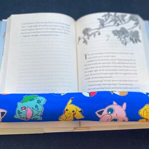 May include: A blue book holder with a Pokemon pattern. The holder has images of Pikachu, Bulbasaur, and Jigglypuff. The holder is on top of an open book with the page number 7 visible.