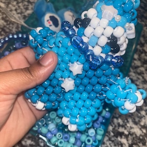 Może przedstawiać: Ręcznie robiona, koralikowa ozdoba w kształcie gwiazdy w odcieniach niebieskiego, białego i czarnego. Ozdoba ma kształt gwiazdy z białymi akcentami w kształcie gwiazd i jest trzymana w dłoni. Ozdoba otoczona jest tacką z koralikami.