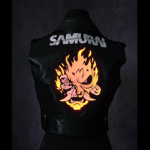 May include: Black leather jacket with the word "SAMURAI" in silver lettering across the shoulders. A vibrant, glowing orange and yellow graphic of a stylized face with flames is centered on the back.